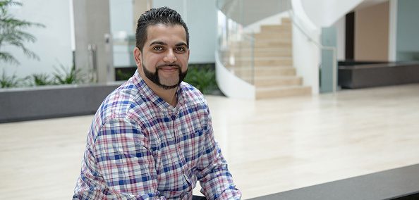 A male employee smiling at the camera, seated indoors in a modern office lobby with a staircase in the background.