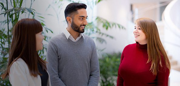 Three colleagues chatting and smiling together in a bright office space with plants.