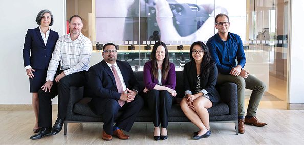 Six coworkers, male & female, posing together in a modern office, some seated on a couch, others standing behind.